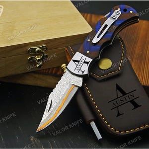 May include: A folding pocket knife with a Damascus steel blade featuring an orange accent line, a handle with blue and brown wood grain patterns, and a silver pocket clip. The blade is engraved with the name AUSTIN, matching the brown leather sheath.