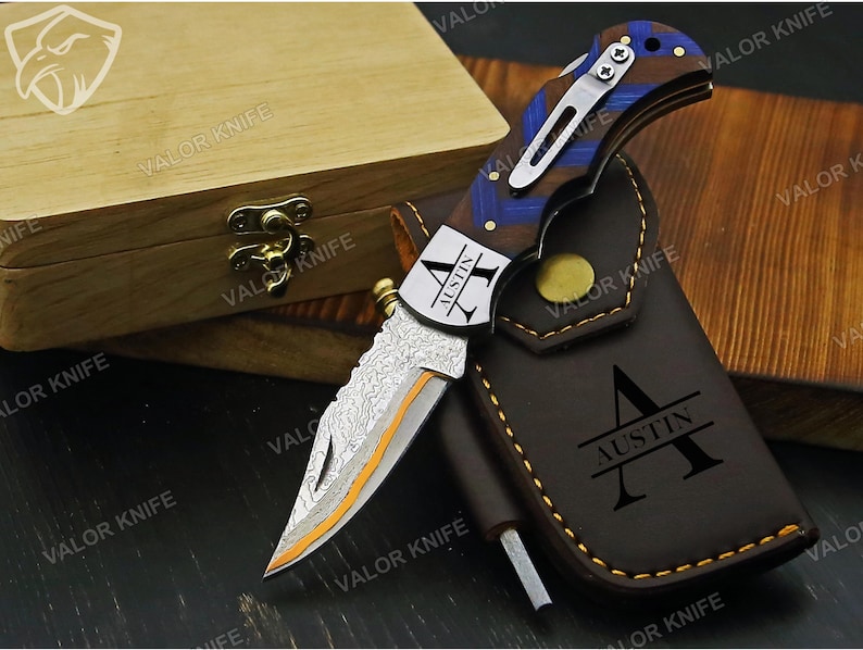 May include: A folding pocket knife with a Damascus steel blade featuring an orange accent line, a handle with blue and brown wood grain patterns, and a silver pocket clip. The blade is engraved with the name AUSTIN, matching the brown leather sheath.