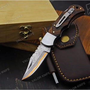 May include: A folding pocket knife with a Damascus steel blade featuring a distinct orange accent line, a dark wood handle with a silver metal pocket clip, and a brown leather sheath with yellow stitching, all branded with the text VALOR KNIFE.