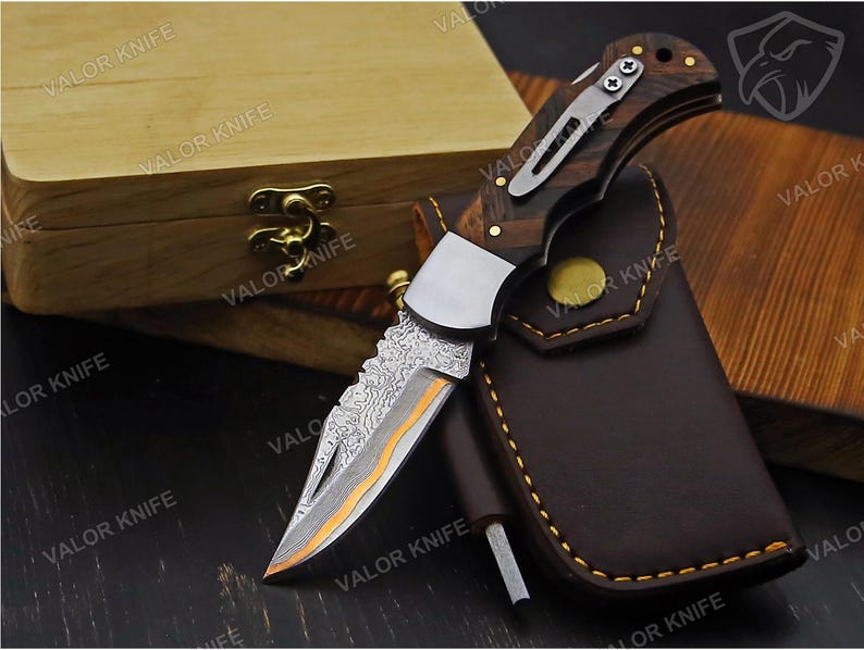 May include: A folding pocket knife with a Damascus steel blade featuring a distinct orange accent line, a dark wood handle with a silver metal pocket clip, and a brown leather sheath with yellow stitching, all branded with the text VALOR KNIFE.