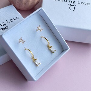 Könnte beinhalten: Zwei Paar goldene Ohrringe mit klaren Zirkonia-Steinen. Die Ohrringe befinden sich in einer weißen Schachtel mit der Aufschrift "Sending Love" auf der Oberseite.