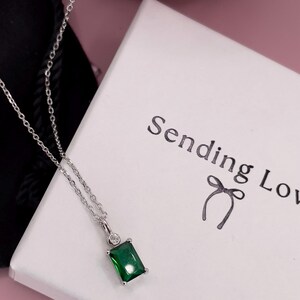 K&ouml;nnte beinhalten: Eine silberne Kettenkette mit einem gr&uuml;nen smaragdartigen Anh&auml;nger und einem kleinen klaren Steinakzent. Die Kette wird auf einer wei&szlig;en Schachtel mit dem aufgedruckten Text "Sending Love" pr&auml;sentiert.