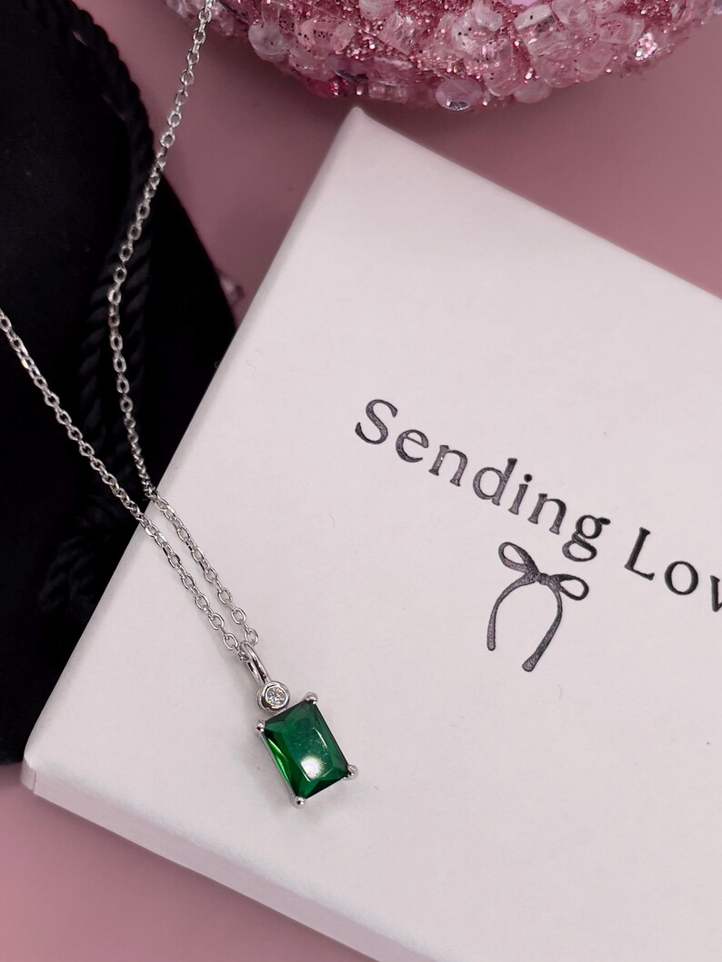 K&ouml;nnte beinhalten: Eine silberne Kettenkette mit einem gr&uuml;nen smaragdartigen Anh&auml;nger und einem kleinen klaren Steinakzent. Die Kette wird auf einer wei&szlig;en Schachtel mit dem aufgedruckten Text "Sending Love" pr&auml;sentiert.