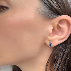 May include: A close-up of an ear with a small, rectangular blue gemstone earring. The earring is a deep blue color and is set in a silver or white metal. The text "Sending Love" is visible.