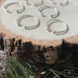 May include: Several open silver rings, each with a small gemstone in either turquoise or pink. The rings are displayed on a natural wooden surface, with a blurred background of green foliage. The rings are a simple, elegant design.