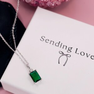K&ouml;nnte beinhalten: Eine silberne Halskette mit einem gr&uuml;nen smaragdartigen Anh&auml;nger und einem kleinen, klaren Stein als Akzent. Die Halskette liegt auf einer wei&szlig;en Schachtel mit dem Text "Sending Love" und einer Schleife.