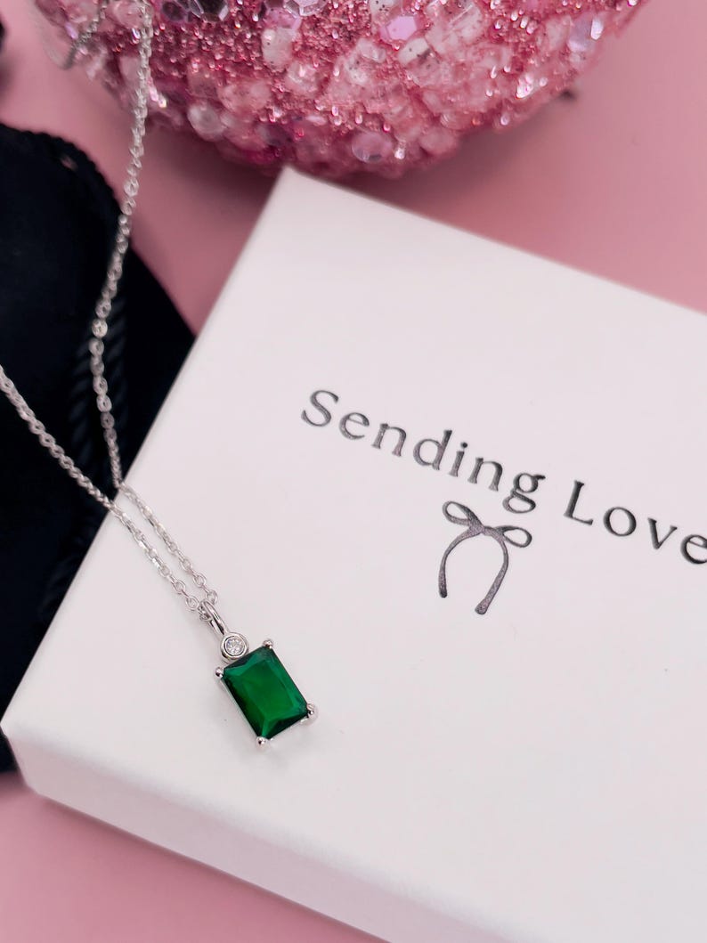 K&ouml;nnte beinhalten: Eine silberne Halskette mit einem gr&uuml;nen smaragdartigen Anh&auml;nger und einem kleinen, klaren Stein als Akzent. Die Halskette liegt auf einer wei&szlig;en Schachtel mit dem Text "Sending Love" und einer Schleife.