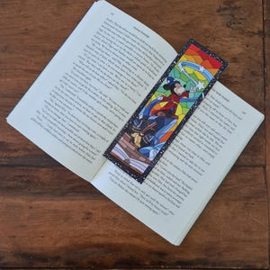 May include: A stained glass bookmark featuring Mickey Mouse in a blue and red outfit, holding a wand with stars. The background is a rainbow of colors with a black border.
