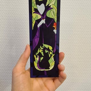 Puede incluir: Un marcap&aacute;ginas de estilo vitral con una silueta de Mal&eacute;fica de La Bella Durmiente de Disney. El marcap&aacute;ginas es morado, verde y amarillo con una silueta negra de Mal&eacute;fica y su drag&oacute;n.