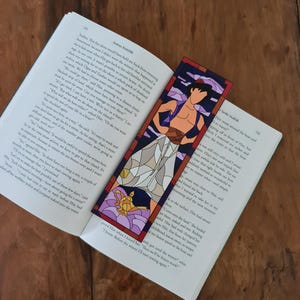 May include: A close-up of a book with a bookmark featuring a stained glass style illustration of Aladdin from the Disney movie Aladdin. The bookmark is purple, blue, and gold. The book is open to a page with text about the movie.