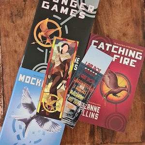 May include: Books from The Hunger Games series by Suzanne Collins are displayed on a wooden surface. The books include "The Hunger Games", "Catching Fire", and "Mockingjay". A bookmark with a stained-glass design and text is also visible.