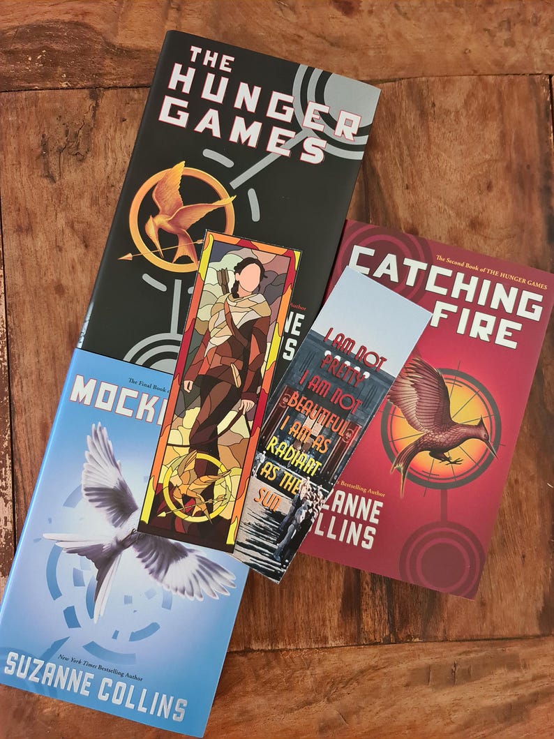 May include: Books from The Hunger Games series by Suzanne Collins are displayed on a wooden surface. The books include "The Hunger Games", "Catching Fire", and "Mockingjay". A bookmark with a stained-glass design and text is also visible.