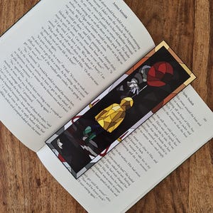 Puede incluir: Un marcapáginas de vidrieras de colores con una figura con una túnica amarilla y globos. El marcapáginas es rectangular con un borde negro y está colocado en un libro abierto.