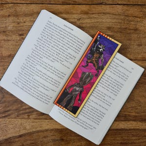 May include: A close-up of a book with a bookmark featuring a stained glass style image of Rocket Raccoon and Groot from the Guardians of the Galaxy. The bookmark is placed on the page with the text "Andrus K" and "Spoke S" visible.