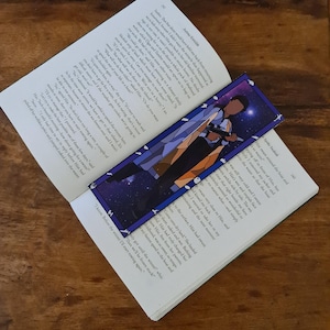 May include: A bookmark featuring a character from a fantasy novel. The bookmark is made of cardstock and has a colourful stained glass design. The character is depicted in a blue and white outfit, holding a sword and standing in front of a starry night sky.