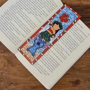 Könnte beinhalten: Ein farbenfrohes Lesezeichen mit einer Zeichentrickfigur aus dem Disney-Film Lilo & Stitch. Das Lesezeichen ist in Form eines Buntglasfensters mit einer roten Hibiskusblüte und einer blauen Figur mit großen Ohren gestaltet. Der Text auf dem Lesezeichen lautet "Spoke Snakish".