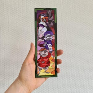 Op de afbeelding: Een glas-in-lood bladwijzer met personages uit de animatiefilm De Reddertjes. De bladwijzer toont een muis in een rode trui, een muis in een paarse sjaal en een pingu&iuml;n in een wit-zwarte hoed.