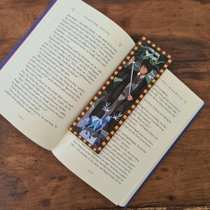 Puede incluir: Un primer plano de un libro con un marcapáginas que presenta una ilustración de vitral de un mago sosteniendo una espada y un ciervo. El título del libro es "Harry Potter y la Cámara Secreta".