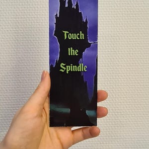 Puede incluir: Un marcap&aacute;ginas morado y negro con una silueta de un castillo y el texto "Touch the Spindle".