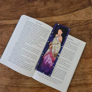 Puede incluir: Un marcapáginas de vitral con una mujer con un vestido blanco sobre un fondo rosa y morado. El marcapáginas es de forma rectangular con un borde azul oscuro.