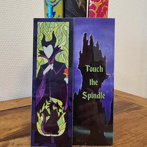 Puede incluir: Dos marcap&aacute;ginas con personajes de la pel&iacute;cula de Disney La Bella Durmiente. El primer marcap&aacute;ginas muestra a Mal&eacute;fica en un estilo de vitral con su forma de drag&oacute;n en el fondo. El segundo marcap&aacute;ginas muestra una silueta del castillo de la Bella Durmiente con el texto "Toca el huso".