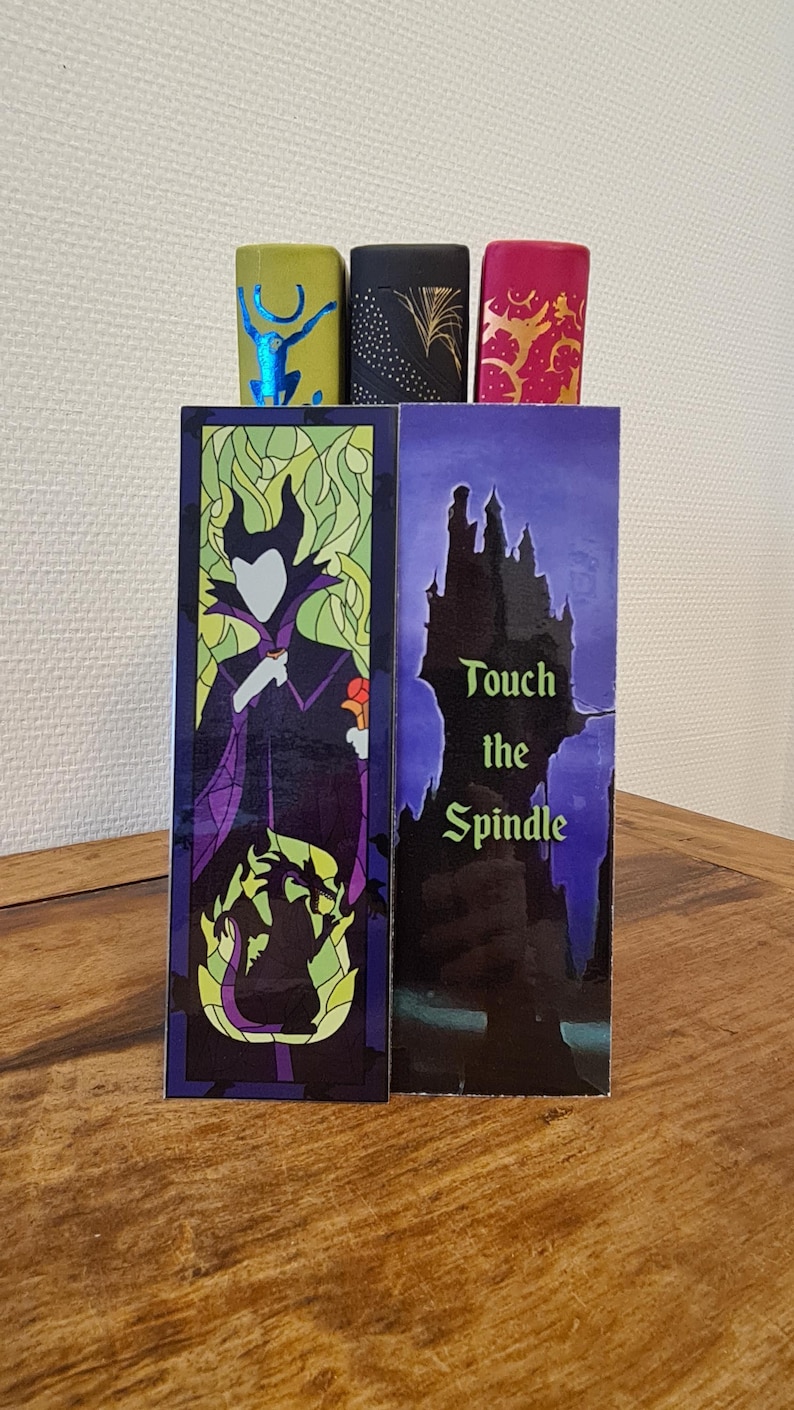 Puede incluir: Dos marcap&aacute;ginas con personajes de la pel&iacute;cula de Disney La Bella Durmiente. El primer marcap&aacute;ginas muestra a Mal&eacute;fica en un estilo de vitral con su forma de drag&oacute;n en el fondo. El segundo marcap&aacute;ginas muestra una silueta del castillo de la Bella Durmiente con el texto "Toca el huso".