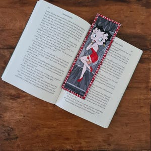 Puede incluir: Un primer plano de un libro con un marcapáginas que presenta un personaje de dibujos animados. El marcapáginas es rojo, blanco y negro con un borde de corazón rojo. El personaje de dibujos animados es una mujer con cabello rojo y un vestido rojo. El libro está abierto en una página con texto sobre un viaje.