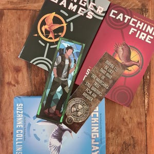 May include: A collection of "The Hunger Games" book series by Suzanne Collins. The books are in green, red, and blue, with the titles "The Hunger Games", "Catching Fire", and "Mockingjay". A bookmark with a character illustration and text is also present.