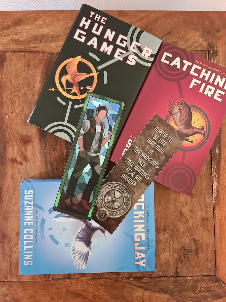 May include: A collection of "The Hunger Games" book series by Suzanne Collins. The books are in green, red, and blue, with the titles "The Hunger Games", "Catching Fire", and "Mockingjay". A bookmark with a character illustration and text is also present.