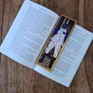 Peut inclure: Un marque-page en vitrail représentant un portrait d'un homme en uniforme blanc et bleu avec un chapeau noir. L'homme tient une tasse dans sa main gauche. Le marque-page est encadré d'une bordure dorée. Le texte "Spoke Smack" est visible dans le coin inférieur droit du marque-page.