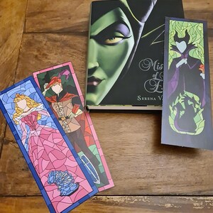 Puede incluir: Tres marcap&aacute;ginas con ilustraciones de personajes de la pel&iacute;cula de Disney "La Bella Durmiente" en estilo de vidriera. Los marcap&aacute;ginas est&aacute;n sobre una superficie de madera. Los marcap&aacute;ginas representan a Aurora, al Pr&iacute;ncipe Felipe y a Mal&eacute;fica.