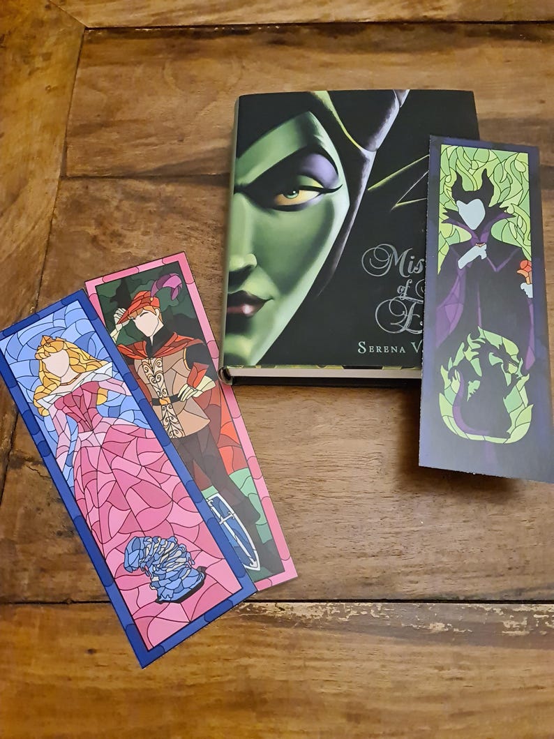 Puede incluir: Tres marcap&aacute;ginas con ilustraciones de personajes de la pel&iacute;cula de Disney "La Bella Durmiente" en estilo de vidriera. Los marcap&aacute;ginas est&aacute;n sobre una superficie de madera. Los marcap&aacute;ginas representan a Aurora, al Pr&iacute;ncipe Felipe y a Mal&eacute;fica.