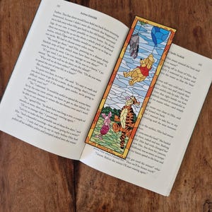 Könnte beinhalten: Ein rechteckiger Lesezeichen mit Buntglas-Design, das Winnie Puuh, Ferkel und Tiger zeigt. Das Lesezeichen liegt auf einem offenen Buch und zeigt eine farbenfrohe Szene.