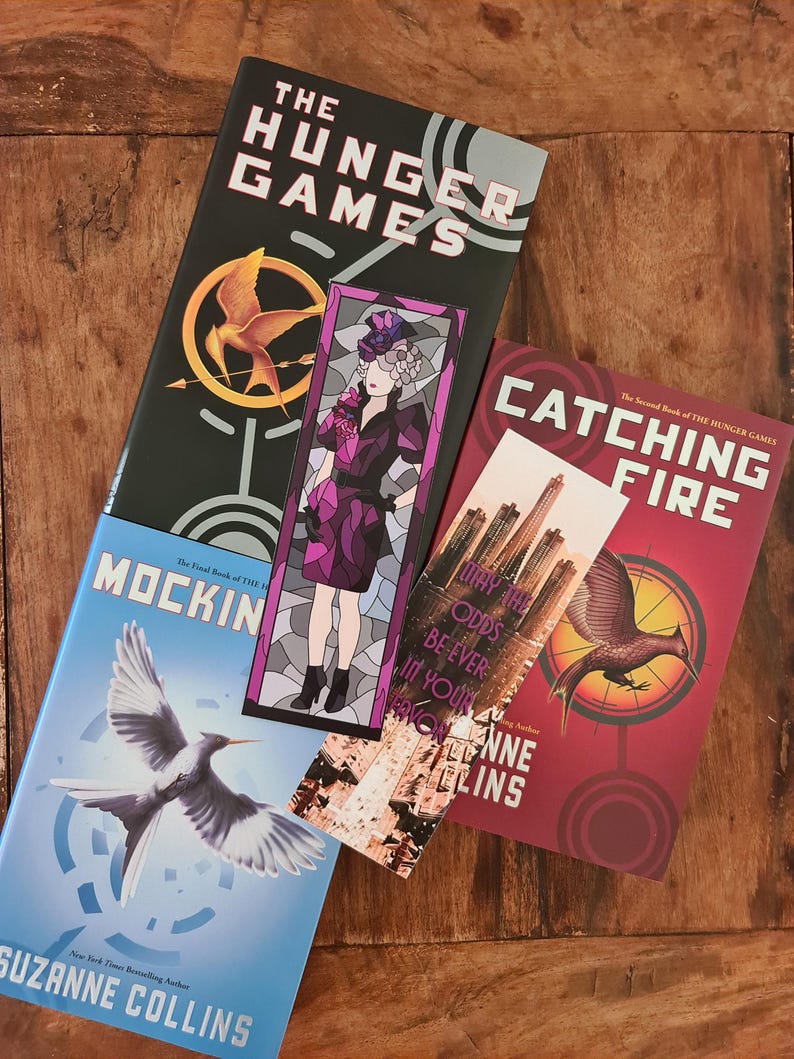 May include: Books from The Hunger Games series, including "The Hunger Games," "Catching Fire," and "Mockingjay." The covers are in black, red, and blue, with images of a mockingjay, a phoenix, and a woman. Bookmarks are also visible.