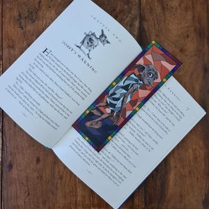 May include: A close-up of an open book with a bookmark featuring a cartoon elf with large ears and a stained glass design. The book's pages are open to a chapter titled "Dobby's Warning" and the text reads "Harry managed not to shout out, but it was a close thing... bulging green eyes the size of tennis balls. Harry knew instantly that this was what had been watching him out of the garden hedge..."