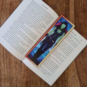 Puede incluir: Un marcapáginas con un personaje de superhéroe verde y morado con una corona de hojas. El personaje está de pie frente a un fondo de vitrales azul y blanco. El marcapáginas se titula "Spoke Snakish".