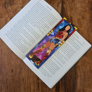 Puede incluir: Un marcapáginas de vitral con Wonder Woman en su icónico traje rojo, azul y dorado. El marcapáginas está sobre un fondo azul y morado.