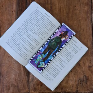 Può includere: Un segnalibro colorato con un'illustrazione in stile vetrata di una persona in abiti verdi e due pesci viola. Il segnalibro è posto in un libro aperto su una superficie di legno.