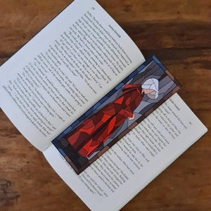May include: A rectangular bookmark featuring a stained-glass style illustration of a person in a red cloak and white head covering. The bookmark is placed on an open book with text visible.