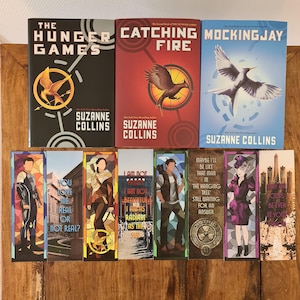 May include: A collection of books and bookmarks from "The Hunger Games" series. The book covers are black, red, and blue, featuring the series title and author's name. Bookmarks display character illustrations and quotes.
