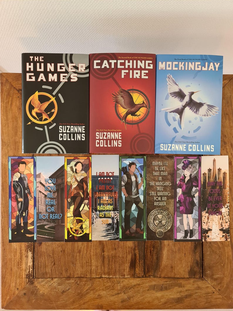 May include: A collection of books and bookmarks from "The Hunger Games" series. The book covers are black, red, and blue, featuring the series title and author's name. Bookmarks display character illustrations and quotes.