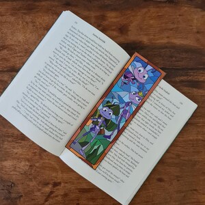 May include: A stained glass bookmark featuring a cartoon ant with a blue body, red eyes, and a green hat. The ant is carrying a small, green, leafy branch. The background is a green and blue stained glass design.