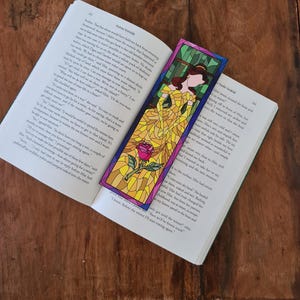 May include: A stained glass bookmark featuring Belle from Disney's Beauty and the Beast. Belle is wearing a yellow dress with a red rose in her hand. The background is a stained glass design of a window with green and blue hues.