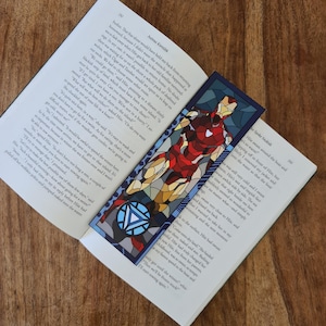 Puede incluir: Primer plano de un libro con un marcapáginas que presenta una imagen de Iron Man en estilo de vitral. El marcapáginas es azul y rojo con un diseño amarillo y dorado. El libro está abierto en una página con texto sobre un viaje por mar.