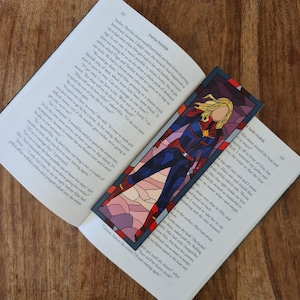 May include: A stained glass bookmark featuring a superhero in a blue and red costume with a star on their chest. The background is a colourful stained glass design with shades of pink, purple, and blue.