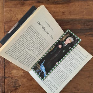 May include: A close-up of a book with a bookmark featuring a cartoon illustration of Harry Potter from the Harry Potter series. The bookmark is green and white with a peacock design. The book is open to a page with the title "The Unbreakable Vow".