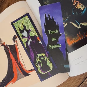 Puede incluir: Dos marcadores de p&aacute;gina con Mal&eacute;fica de la pel&iacute;cula de Disney La Bella Durmiente. Un marcador es un dise&ntilde;o de vitral con Mal&eacute;fica con un vestido morado y cuernos verdes. El otro marcador es una silueta negra del castillo de Mal&eacute;fica con el texto "Touch the Spindle".