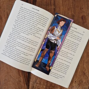 May include: A stained glass bookmark featuring a man in a blue and purple tunic with a gold chain necklace. The bookmark is set against a blue and purple background.