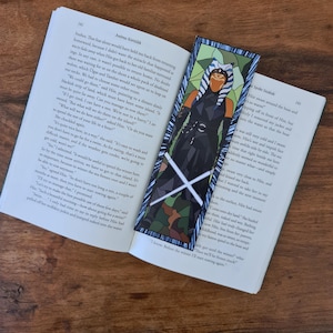 以下が含まれることがあります： SFキャラクターのブックマーク。白黒のデザインで、ライトセーバーが2本描かれています。ブックマークは、テキストが見える開いた本の中にあります。