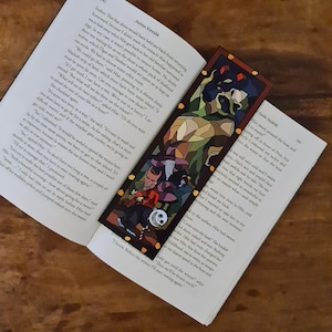 Puede incluir: Primer plano de un marcapáginas con una ilustración de estilo vitral de personajes de la película "Pesadilla antes de Navidad". El marcapáginas está hecho de cartulina gruesa y tiene un borde rojo.